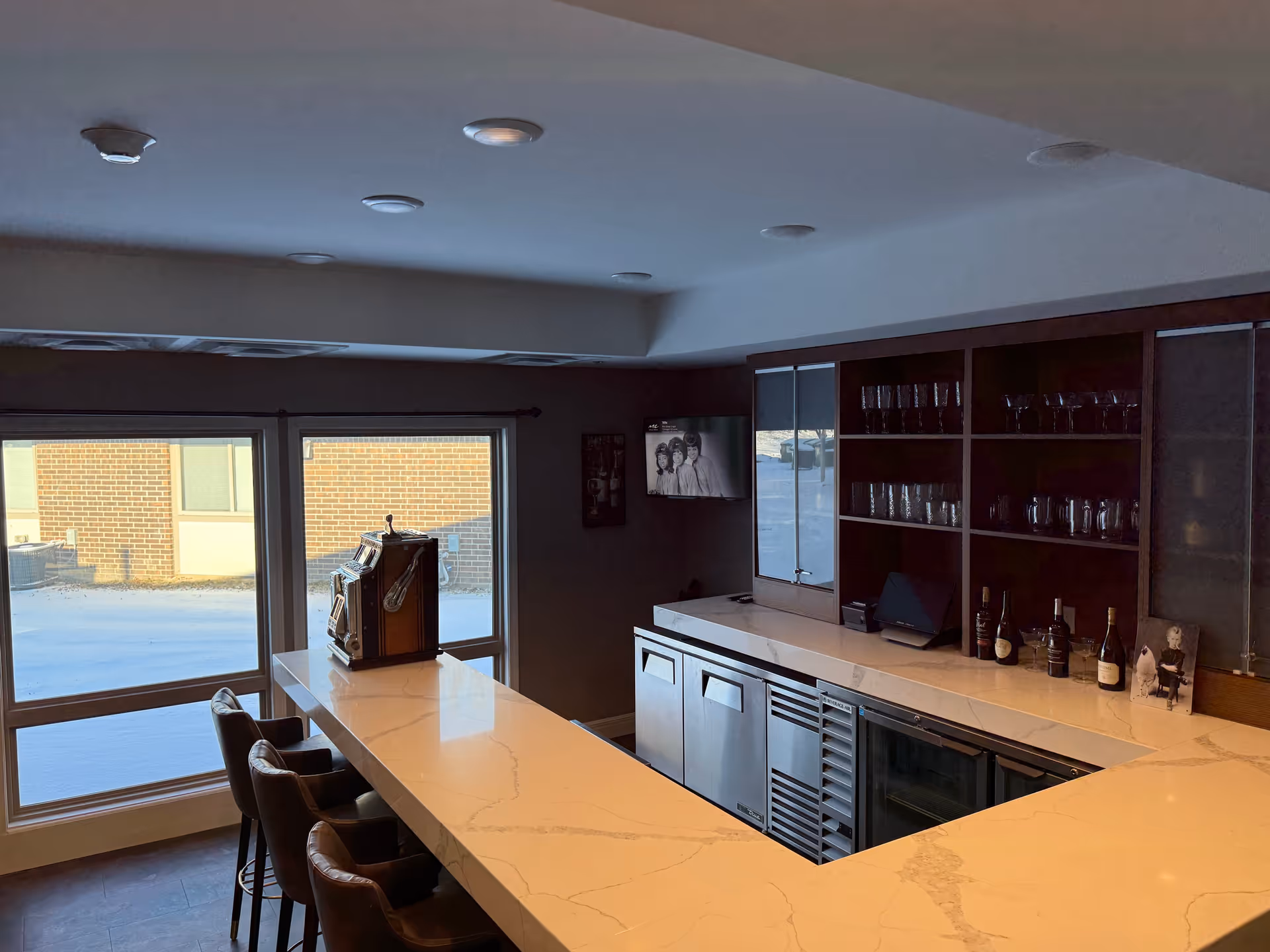 Interior view of a modern bar area with a marble countertop, several bar stools, glass shelves holding glasses, wine bottles on the counter, and a vintage slot machine on the bar. Large windows show a snowy outdoor scene.