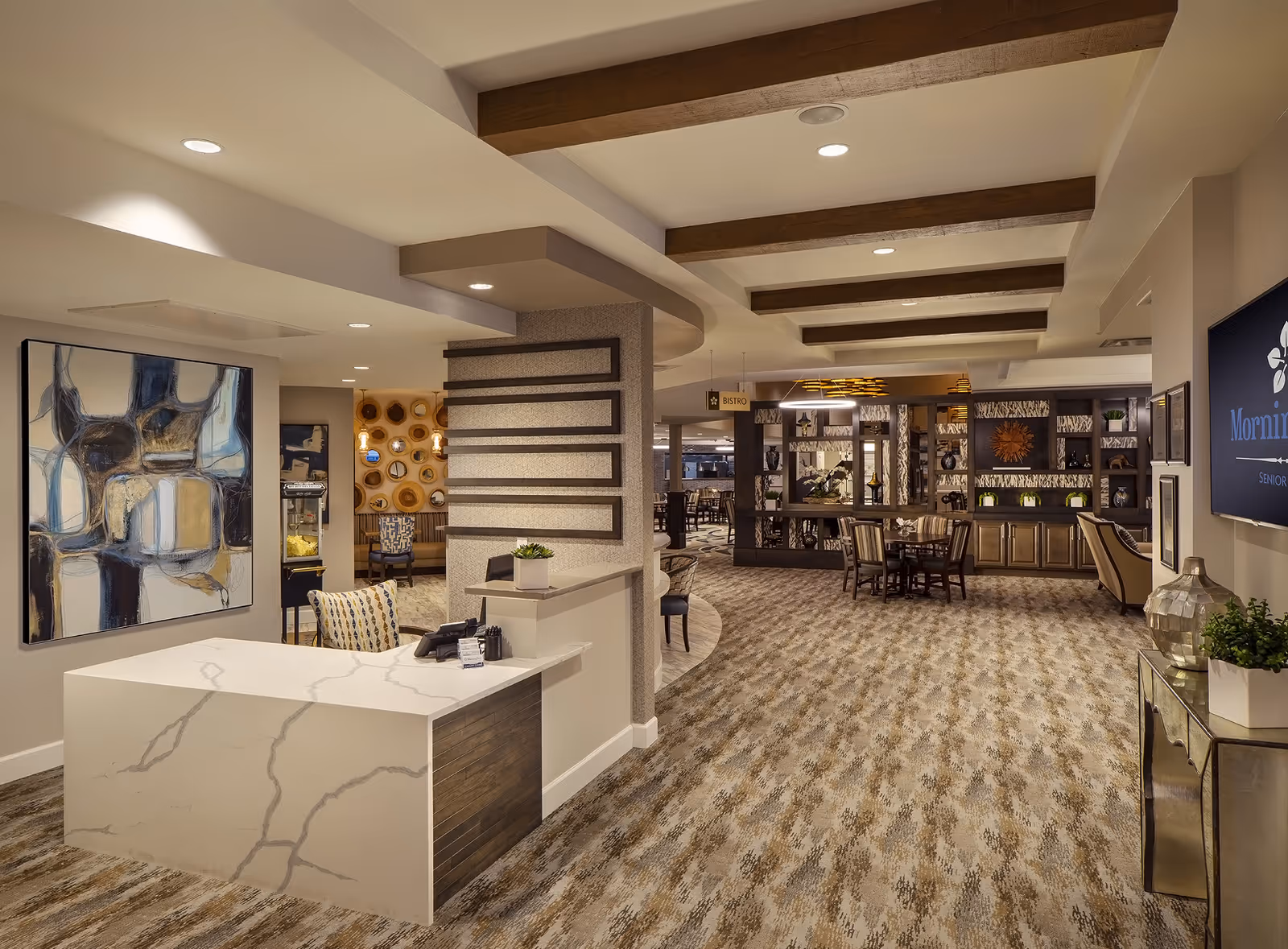 Interior view of a senior living facility featuring a reception desk with a marble countertop and a patterned chair. The space has carpeted floors, wooden ceiling beams, and modern lighting. There is abstract artwork on the wall to the left and a sign indicating a bistro area further inside. The room includes tables and chairs for dining or socializing, decorative shelving, and a television screen on the right displaying the facility name.