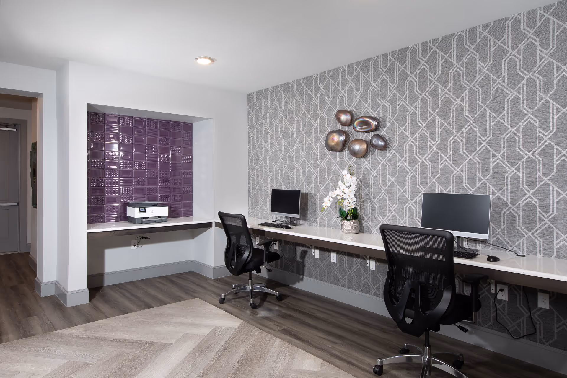 A modern office or computer workstation area with two black mesh office chairs, two desktop computers on a long white desk, a decorative vase with white flowers, and a wall with geometric patterned wallpaper. There is also a recessed area with purple textured tiles and a printer on a small counter. The floor has wood and patterned flooring.