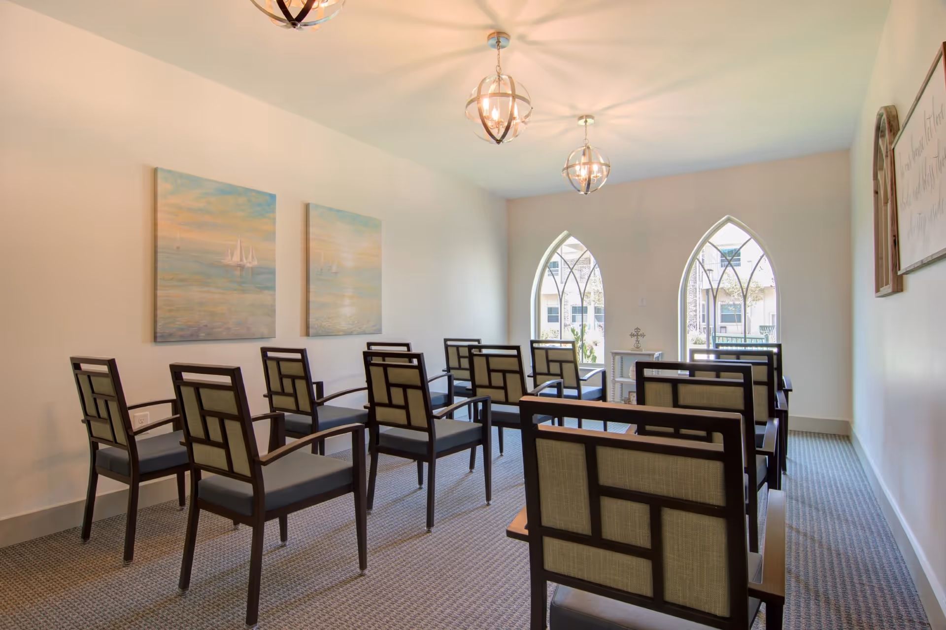 Bright interior room with rows of wooden-framed chairs facing arched windows and wall artwork.