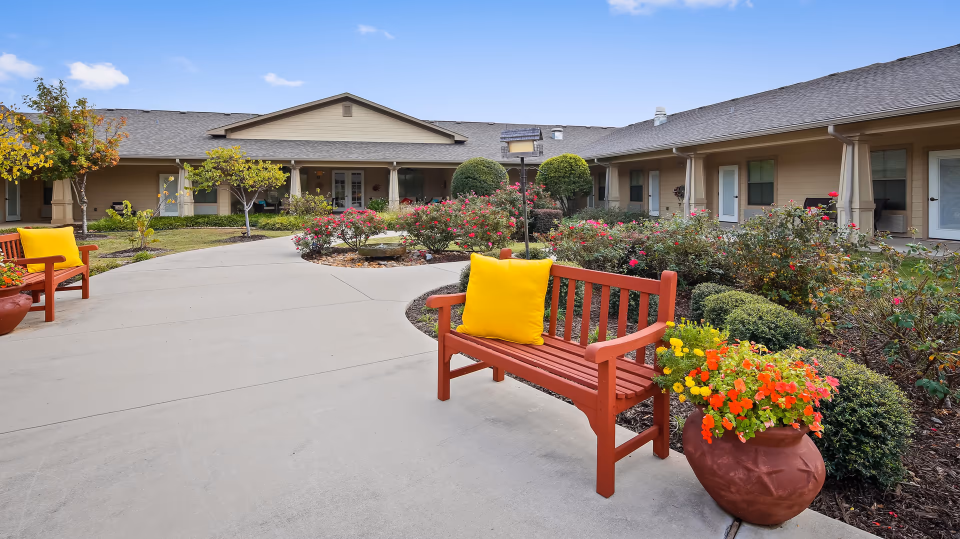 Outdoor courtyard area of an assisted living facility with a concrete pathway, red wooden benches with yellow cushions, potted colorful flowers, and well-maintained bushes and rose plants. The building surrounds the courtyard with beige walls and multiple doors and windows under a gray shingled roof.