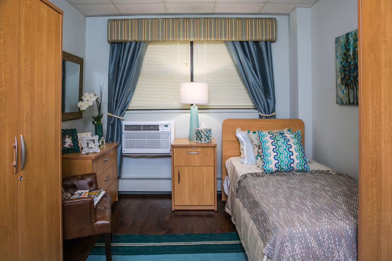 Small furnished single bedroom with a twin bed, nightstand, dresser, armchair, and a window air conditioner.