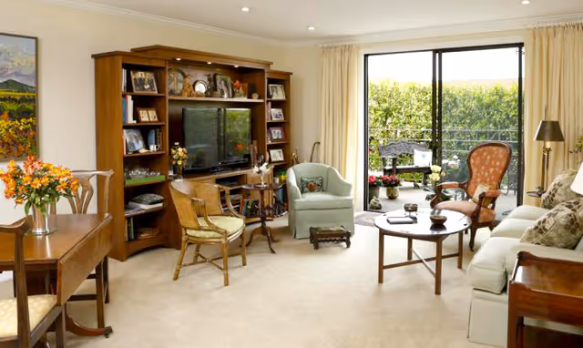 Sunlit living room with sofas and chairs, a wooden entertainment center and coffee table, and sliding glass doors opening to a patio.