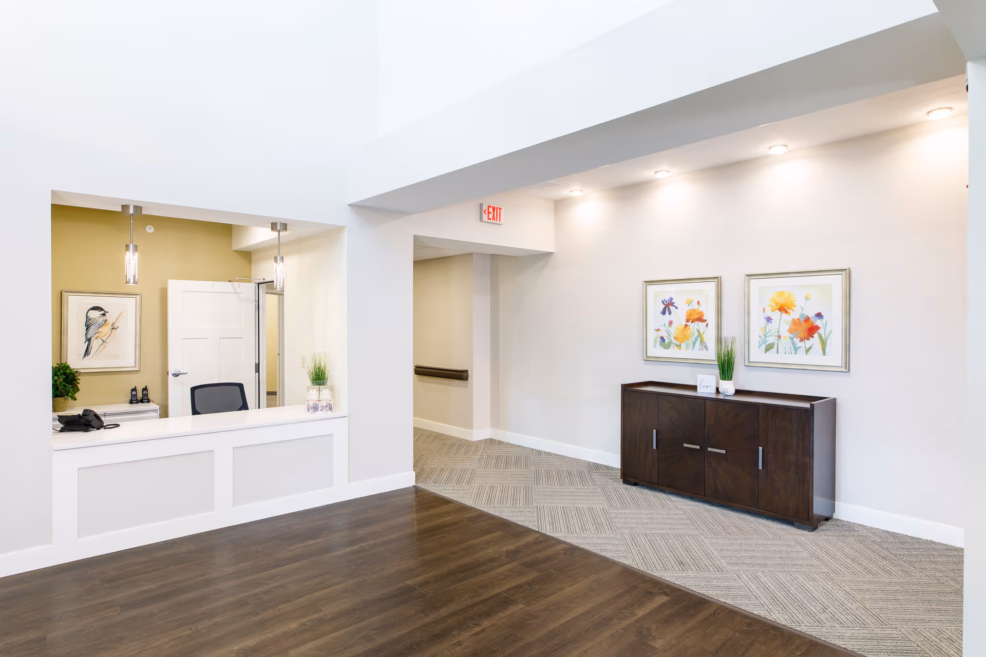 Bright and clean reception area with a white counter and two hanging pendant lights. Behind the counter is a chair, a phone, and a framed bird artwork on a light green wall. To the right, there is a hallway with a dark wooden cabinet topped with a small plant and two framed floral paintings above it. The floor transitions from dark wood to patterned carpet, and there is an illuminated exit sign above the hallway entrance.