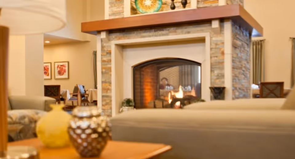 Cozy living room area with a stone fireplace in the center, a fire burning inside, and a blurred view of a person sitting behind the fireplace. The room features comfortable seating, decorative vases on a wooden table in the foreground, and framed artwork on the walls in the background.