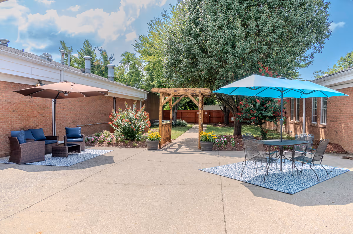 Outdoor patio area at Lebanon Center for Rehabilitation and Healing featuring two seating areas with umbrellas. One side has a brown wicker sofa and chairs with blue cushions under a brown umbrella, and the other side has a metal table and chairs under a blue umbrella. There is a wooden pergola in the center with a pathway leading to a garden area with trees and flowers, surrounded by brick building walls.