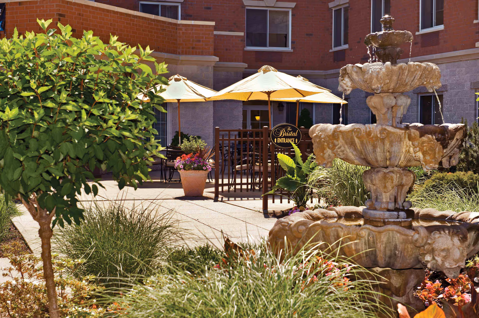 A landscaped courtyard featuring a multi-tiered stone fountain, patio umbrellas, potted flowers and a sign reading 'The Bristal Entrance' in front of a brick building.