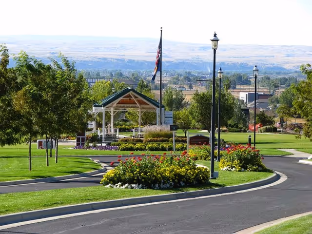 Landscaped outdoor grounds with a white gazebo, an American flag on a pole, lamp posts, a curved paved drive, and colorful flowerbeds with hills in the background.