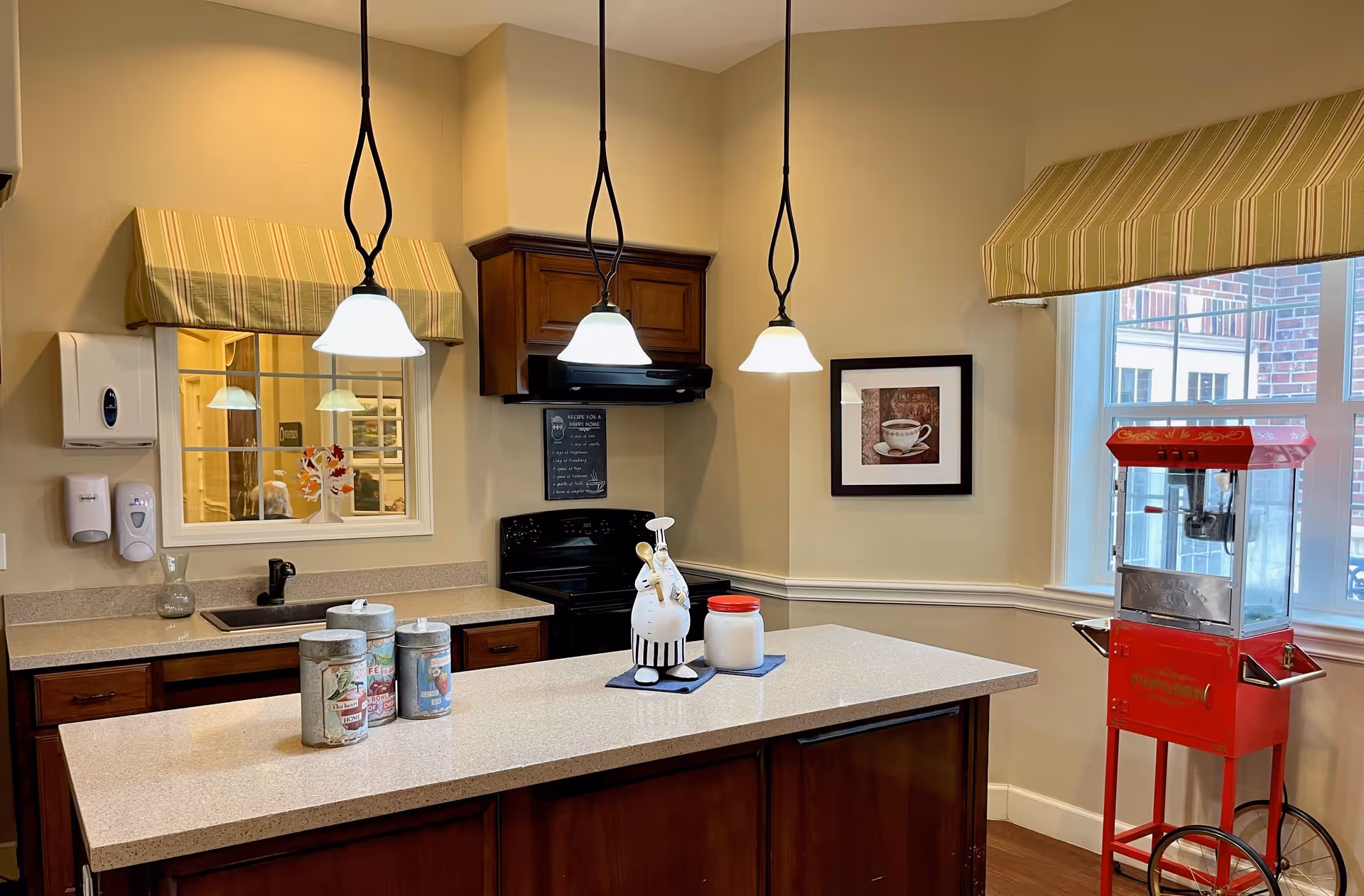Interior view of a kitchen area in a senior living facility with a central island countertop. The kitchen features wooden cabinets, a black stove, a sink, and three hanging pendant lights. On the countertop, there are decorative containers and a ceramic chef figurine. A red popcorn machine is positioned near a window with striped valances. The walls are painted beige, and there is a framed picture of a coffee cup on the wall.