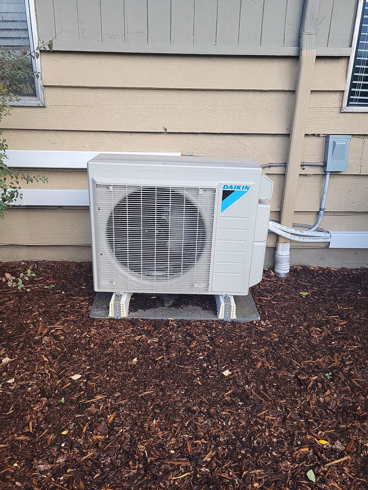 Outdoor air conditioning unit installed on the ground next to the exterior wall of a building with beige siding, surrounded by mulch and some small plants.