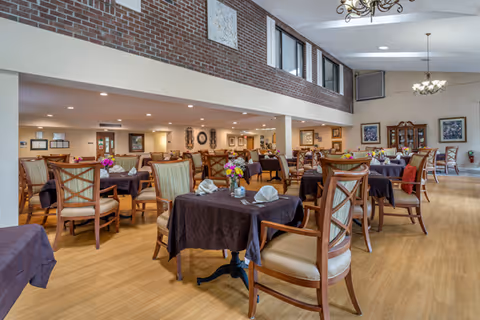 Spacious dining room with multiple tables set with tablecloths, chairs, and floral centerpieces in a senior living facility.