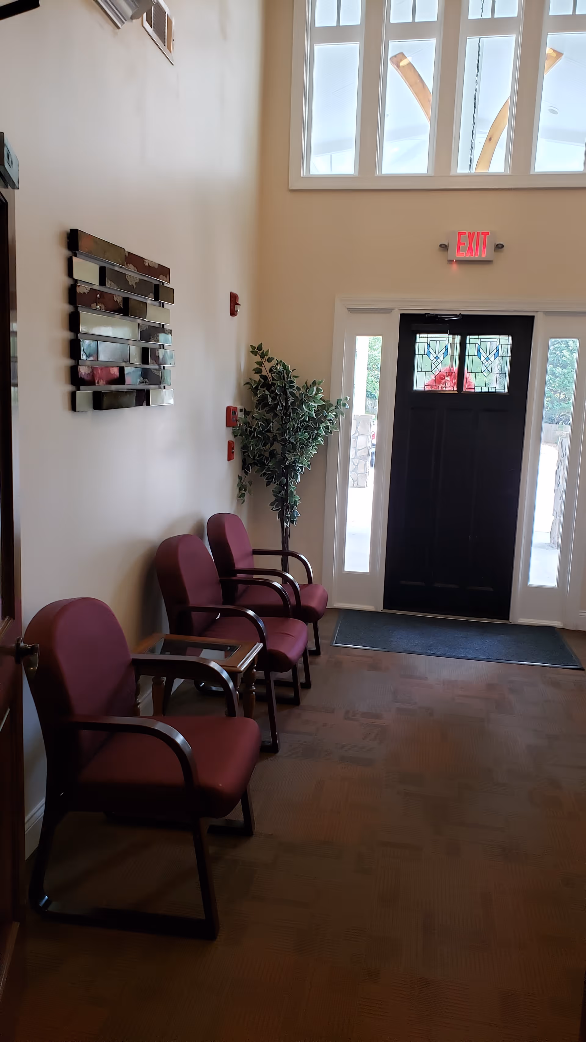 A small waiting area with three maroon cushioned chairs with black armrests arranged along a beige wall. A small wooden side table is placed between two of the chairs. There is a decorative wall mirror with horizontal rectangular segments above the chairs. A tall artificial plant is positioned near a black door with stained glass panels and sidelights. Above the door is a red illuminated exit sign. The floor is carpeted in a muted pattern, and natural light comes through the upper windows.