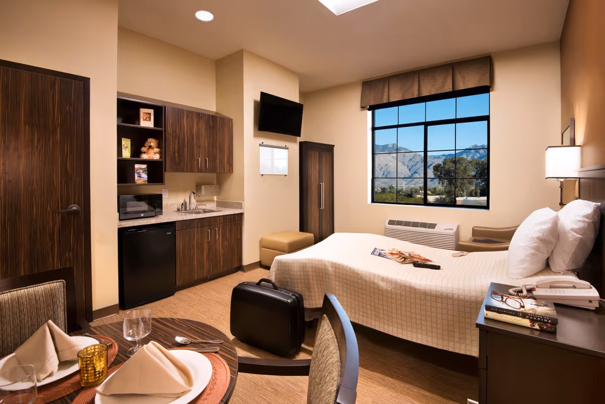A cozy bedroom in a senior living facility featuring a bed with white pillows and a beige bedspread, a bedside table with books, a telephone, and reading glasses. There is a large window with a view of mountains and trees outside. The room includes a small kitchenette with a sink, microwave, mini fridge, and cabinets. A round dining table with two place settings and folded napkins is in the foreground. A flat-screen TV is mounted on the wall above a small ottoman, and a suitcase is placed near the bed.