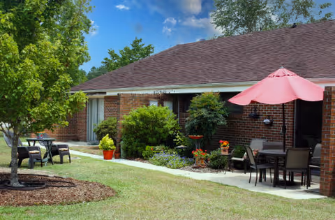 Outdoor patio area of a brick building with a brown roof, featuring a table with a pink umbrella and chairs, another small table with chairs under a tree, and various green shrubs and plants along the walkway.