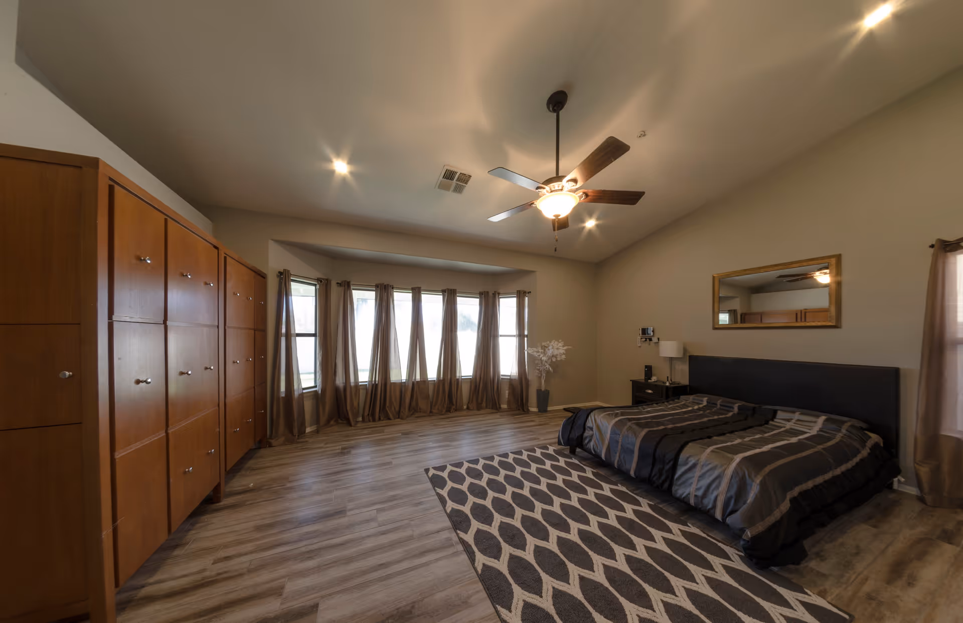 A spacious bedroom with a large bed covered in a dark striped comforter, a patterned area rug, a wooden wardrobe along one wall, a ceiling fan with lights, and a bay window with brown curtains letting in natural light.