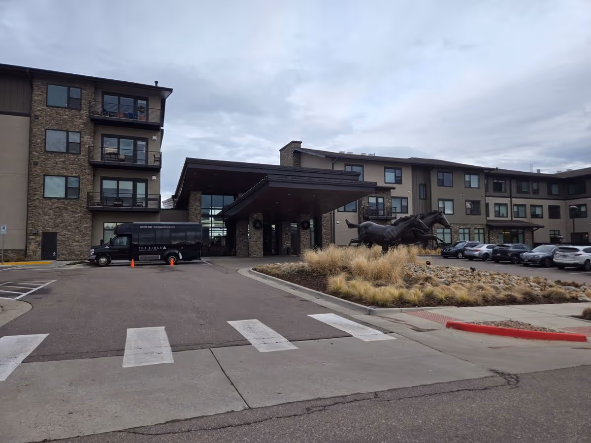 Front entrance of a multi-story senior living building with a covered porte-cochere, a parked shuttle van, and a horse sculpture in the landscaped driveway.