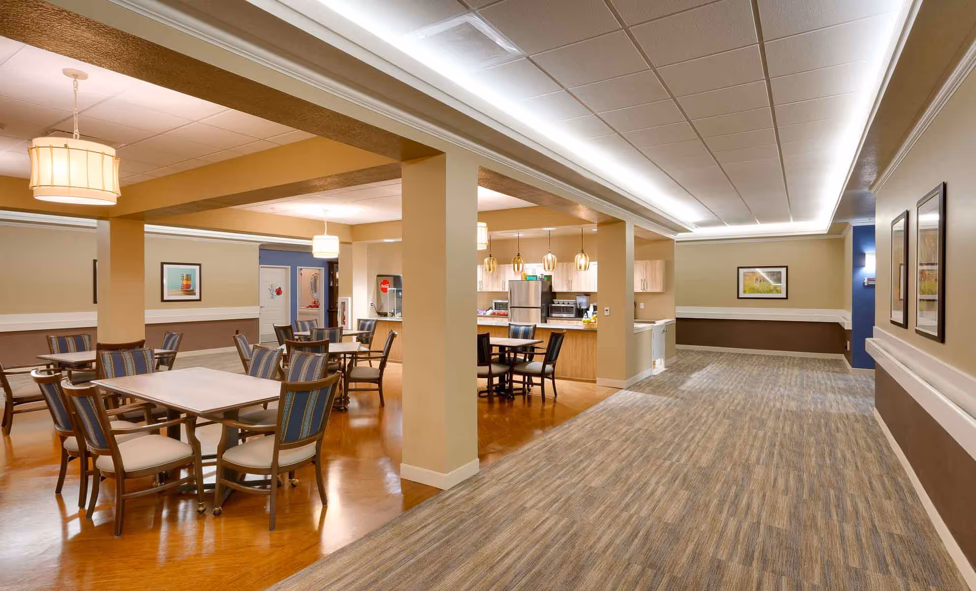 Interior view of a senior living facility dining area with multiple tables and chairs arranged on a wooden floor. The space is well-lit with hanging pendant lights and ceiling lights. In the background, there is a kitchen area with stainless steel appliances and a Coca-Cola vending machine. The walls are painted in neutral tones with framed artwork, and the floor transitions from wood to carpet in the hallway.