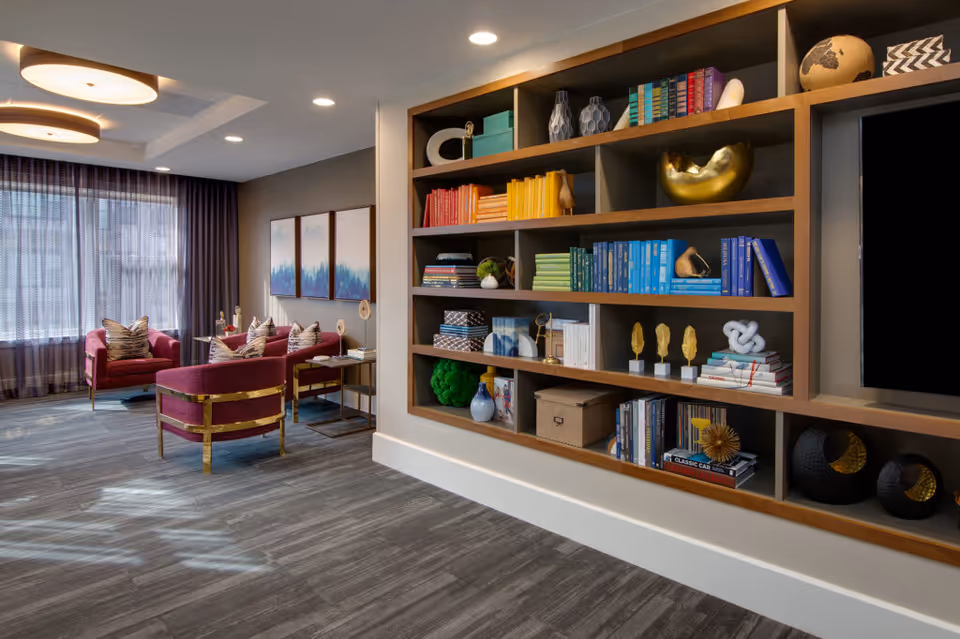 A modern and cozy living room area with a large built-in bookshelf filled with colorful books and decorative items. The room features two red upholstered chairs with patterned pillows, a small side table, and large windows with sheer curtains allowing natural light to enter. The flooring is a gray wood-like material and the ceiling has recessed lighting and circular light fixtures.