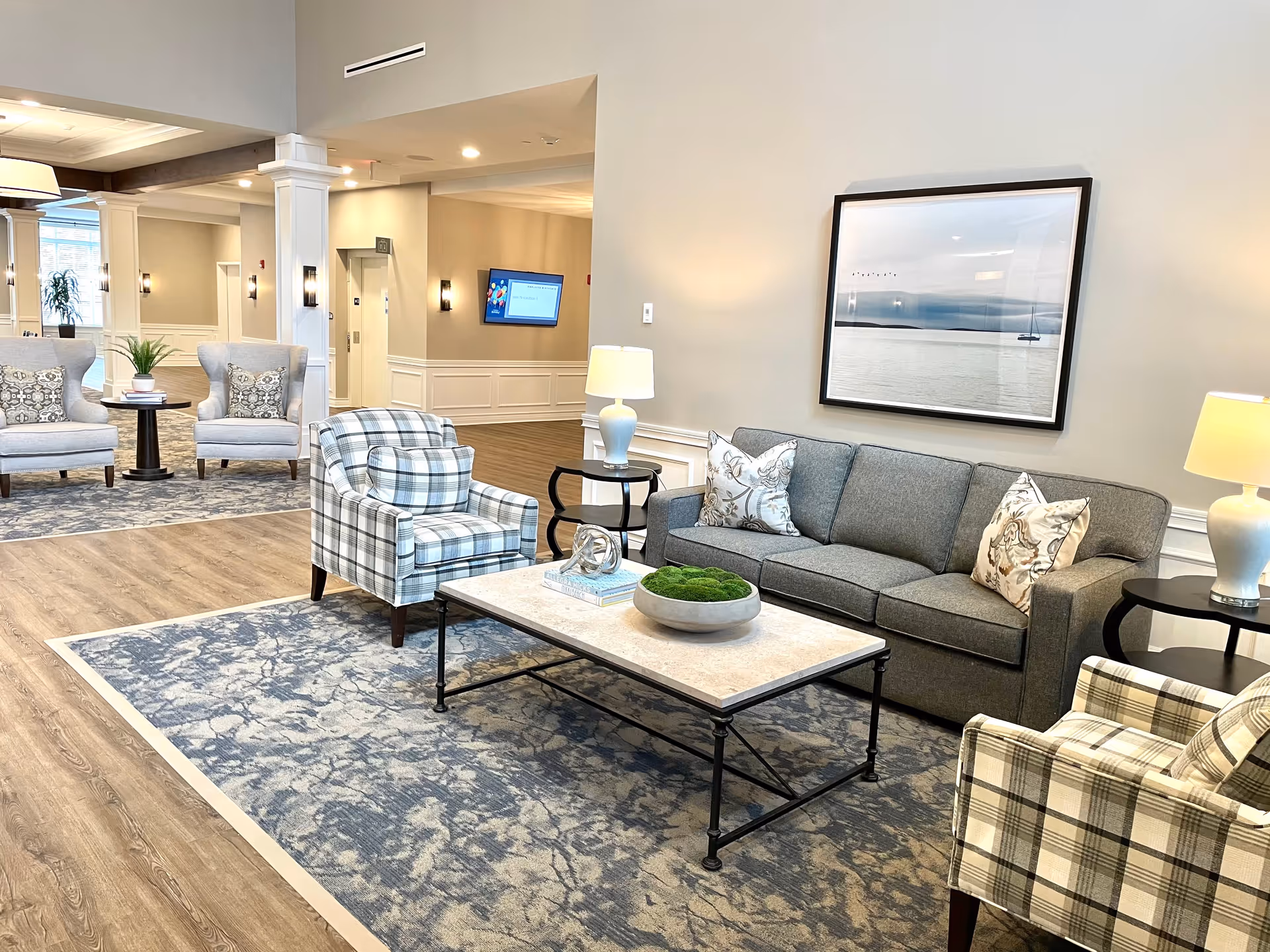 A bright, modern lobby seating area with a gray sofa, plaid armchairs, coffee table, lamps, and wall art.
