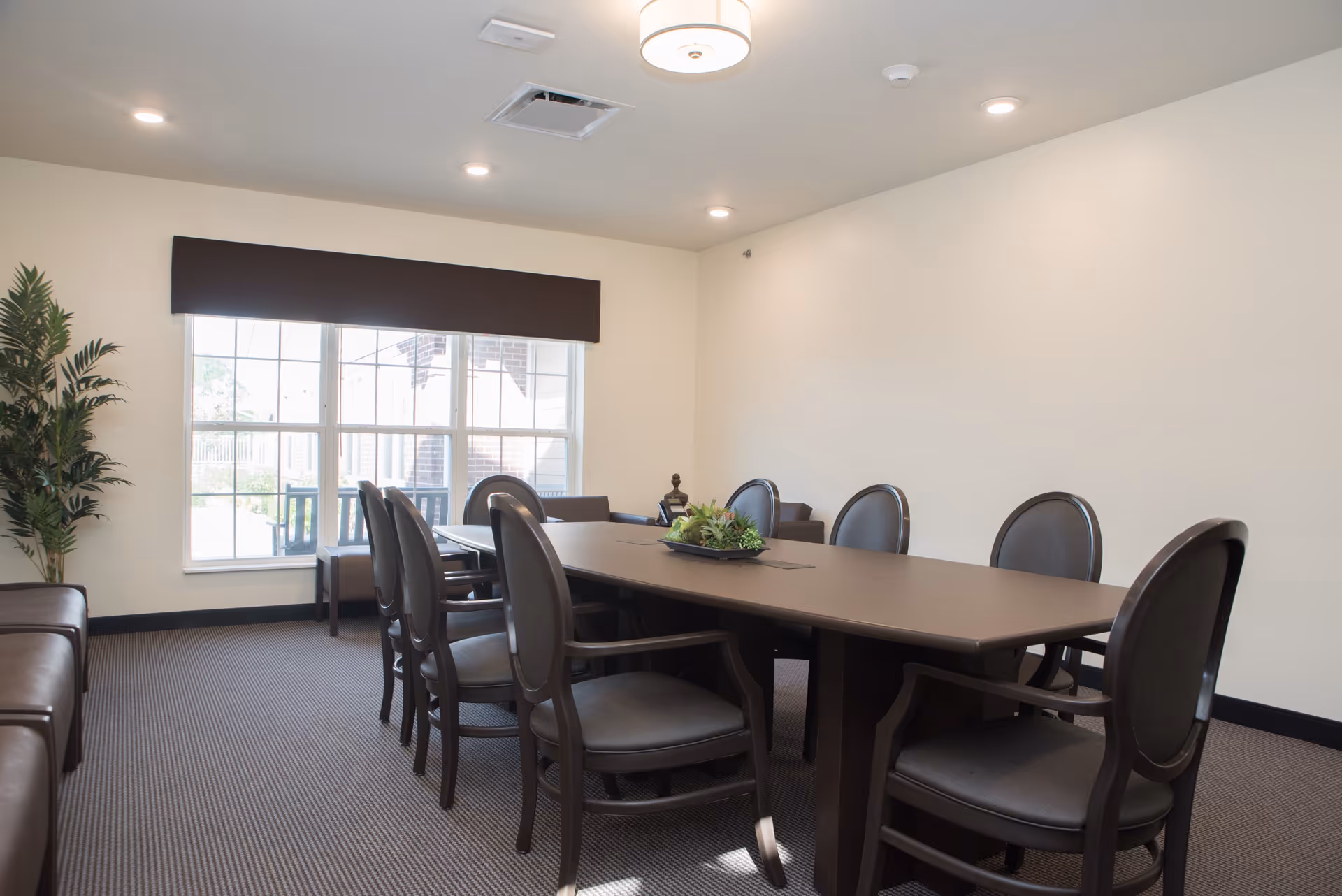 Well-lit meeting/dining room with a long table surrounded by chairs, a large window with a valance, and a potted plant.