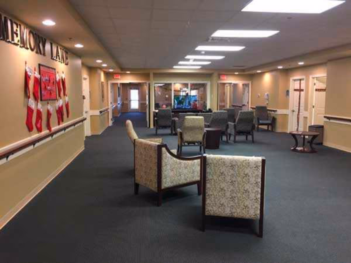 A spacious common area in a senior living facility with several upholstered chairs arranged around small tables. The walls are beige with handrails, and there is a bulletin board decorated with Christmas stockings labeled 'Memory Lane'. The ceiling has recessed lighting and fluorescent panels. Doors and hallways lead to other parts of the facility.