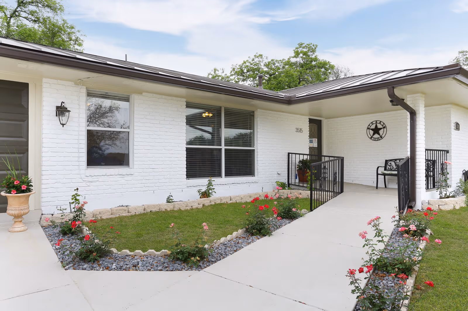 Exterior view of a single-story white brick building with a brown roof, featuring a wheelchair-accessible ramp leading to a covered porch with a black metal railing and a bench. The front yard has a small garden with green grass, red and pink flowers, and decorative rocks along the edges of the walkway.