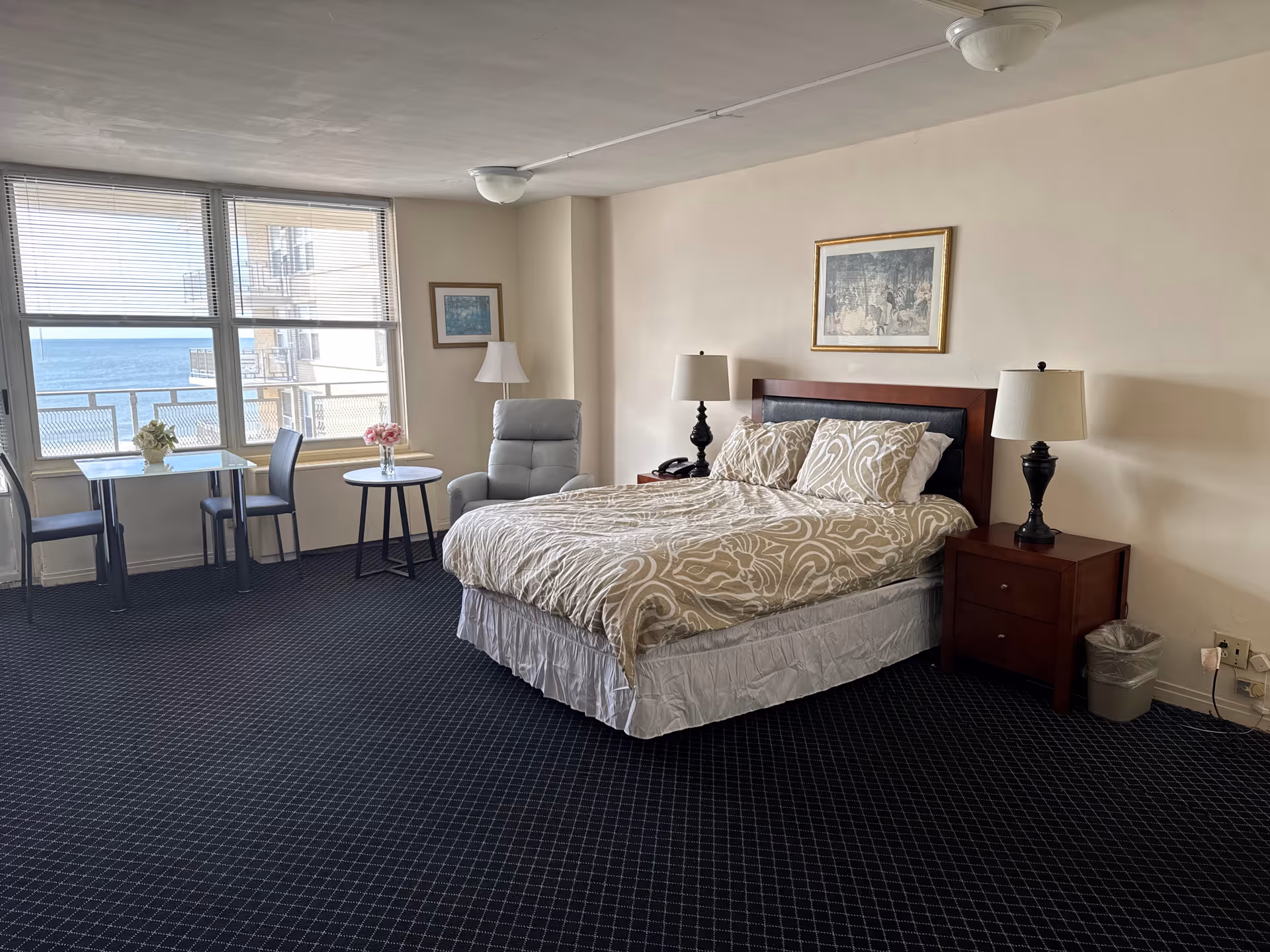 A spacious bedroom with a large bed covered in beige and white patterned bedding, two bedside tables with lamps, a gray armchair, a small round table with a vase of flowers, and a rectangular table with two chairs near a large window showing a view of the ocean and a neighboring building.