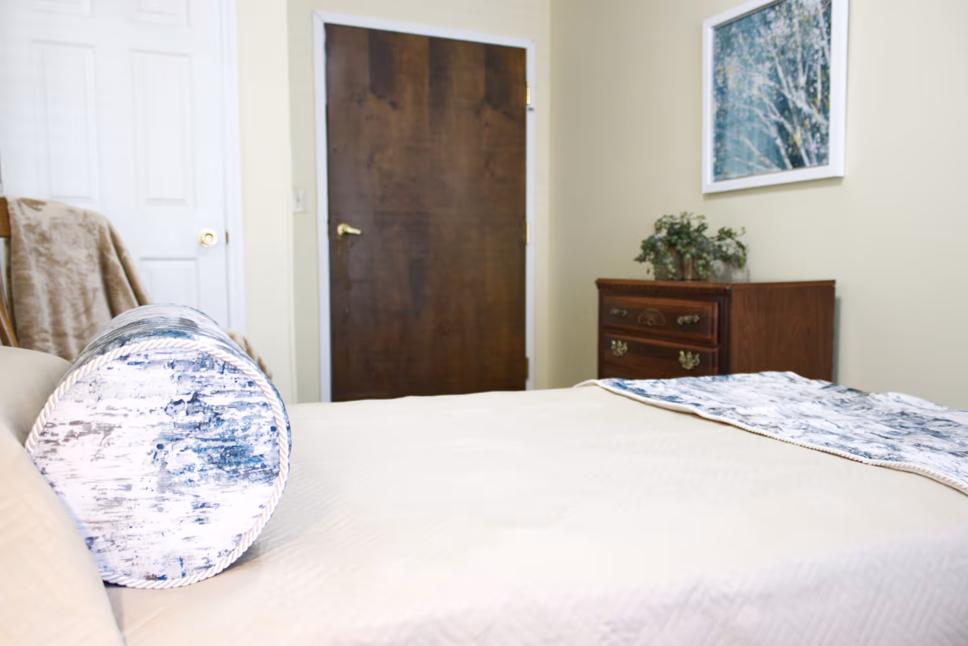 A bedroom with a neatly made bed featuring a cylindrical pillow and a patterned throw blanket. In the background, there is a wooden dresser with a small plant on top, a framed picture on the wall, and two closed doors, one white and one dark brown.