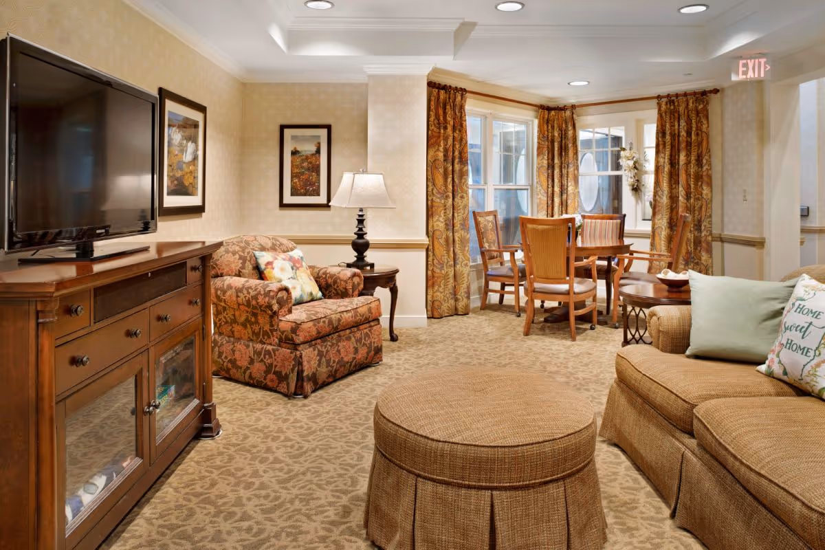 Cozy common room with upholstered seating, ottoman, TV cabinet, and a small dining table by bay windows.