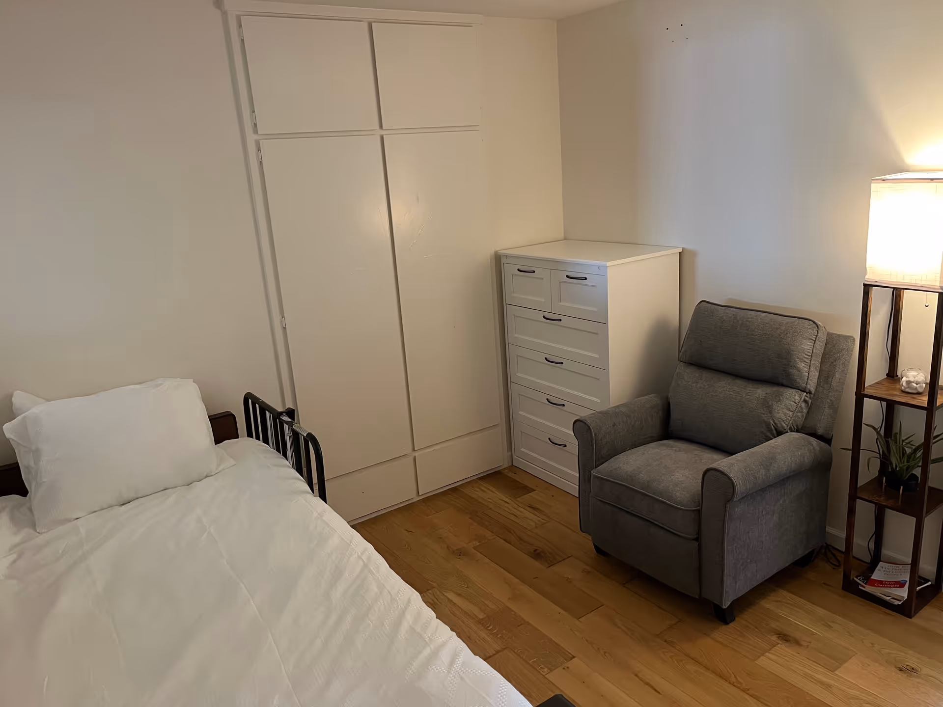 Small bedroom with a single bed, white dresser, gray recliner chair, closet doors and a floor lamp on hardwood floors.