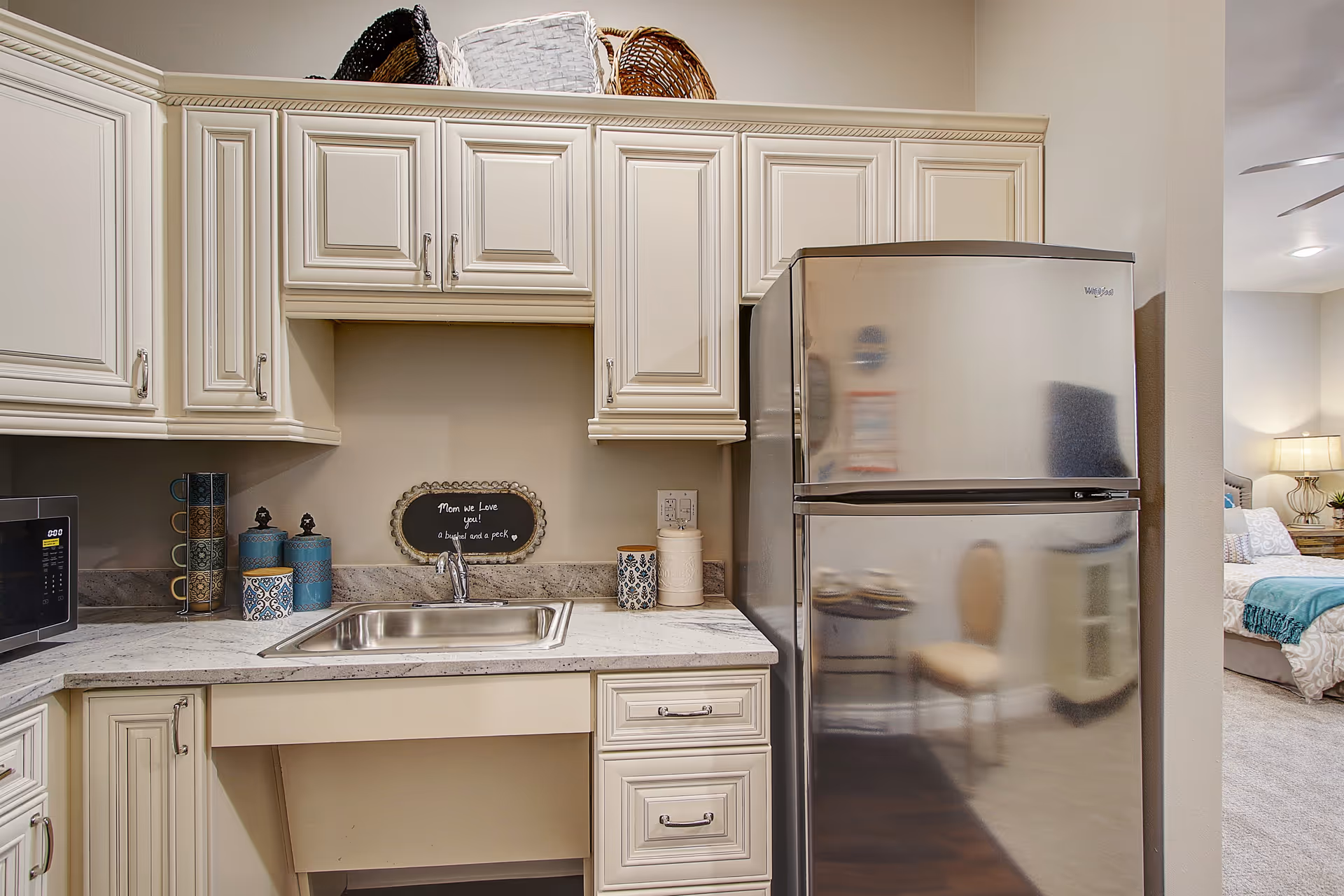 Small kitchenette with cream cabinets, marble countertop, sink, microwave and a stainless-steel refrigerator next to an open bedroom.