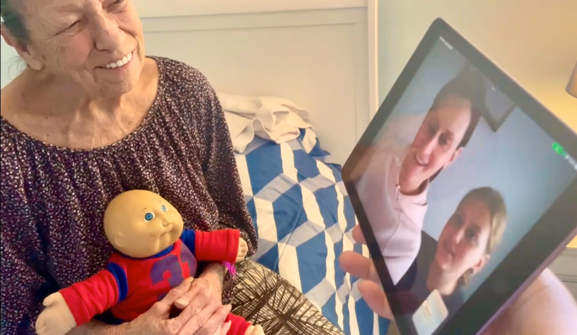 An elderly woman sitting on a bed holding a doll, smiling while looking at a tablet screen showing two younger women on a video call.