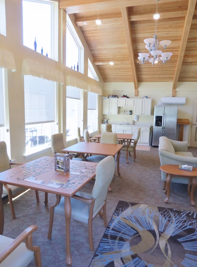 Bright common/dining area with tables and chairs, a jigsaw puzzle on a table, a kitchenette and refrigerator under a vaulted wood ceiling.