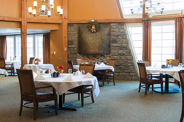 A dining room with several tables covered with white tablecloths, each set with napkins, glasses, and small flower arrangements. The room features large windows with curtains, a stone fireplace, and chandeliers hanging from the ceiling.