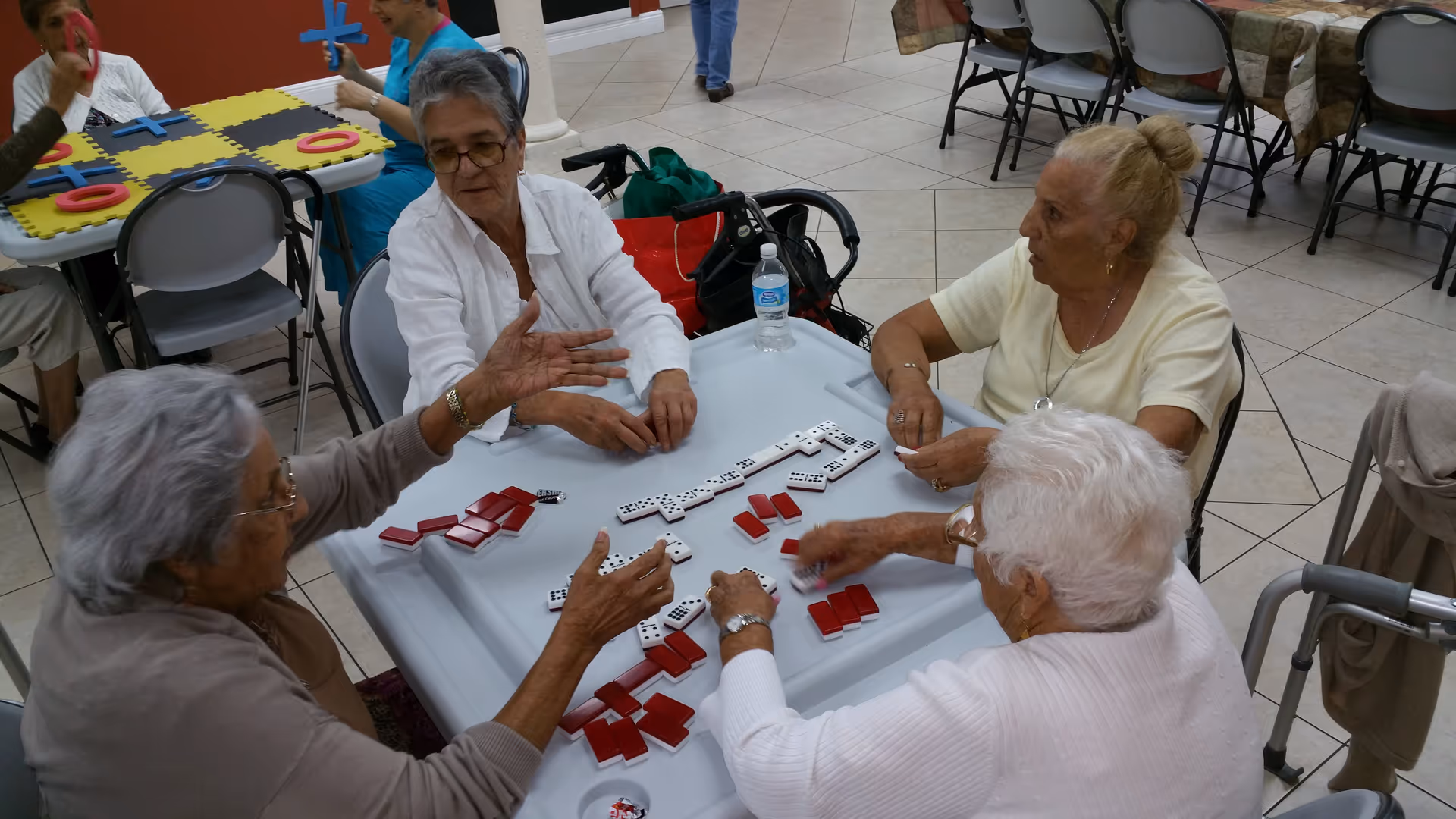 Four elderly women sitting around a table playing a game with dominoes and red tiles in a communal indoor space with tiled floors and other tables and chairs in the background.
