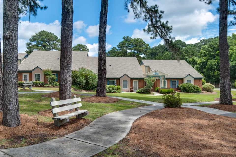 Exterior view of Savannah Square showing a paved walkway, benches and pine trees in front of single-story brick buildings under a blue sky.