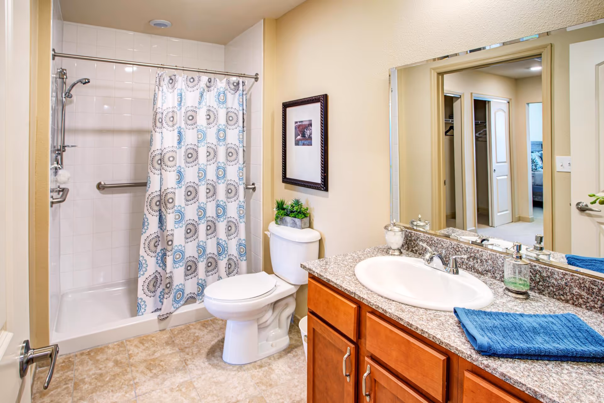 A clean bathroom with a shower and bathtub combination featuring a white and blue patterned shower curtain. There is a white toilet next to the shower, a granite countertop with a white sink, a blue towel, and a small plant. A large mirror above the sink reflects the hallway and adjacent rooms.