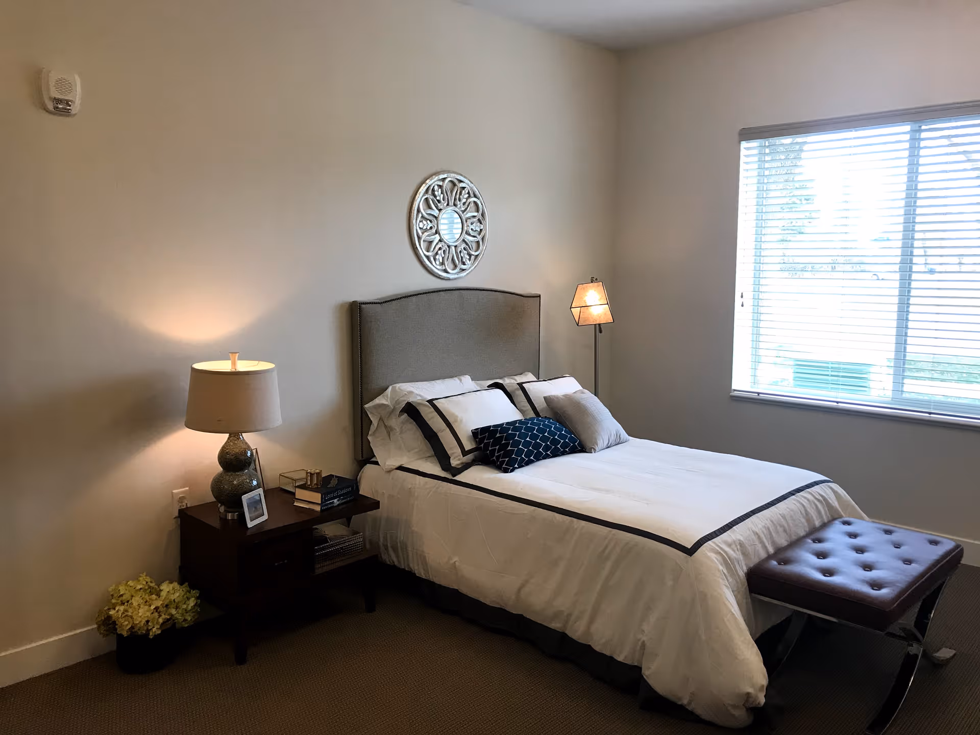 A neatly made bed with white and dark-trimmed bedding in a bedroom. There is a nightstand with a lamp, books, and a photo frame on the left side of the bed. A decorative round mirror hangs on the wall above the headboard. A window with blinds is on the right side, letting in natural light. A cushioned bench is placed at the foot of the bed.