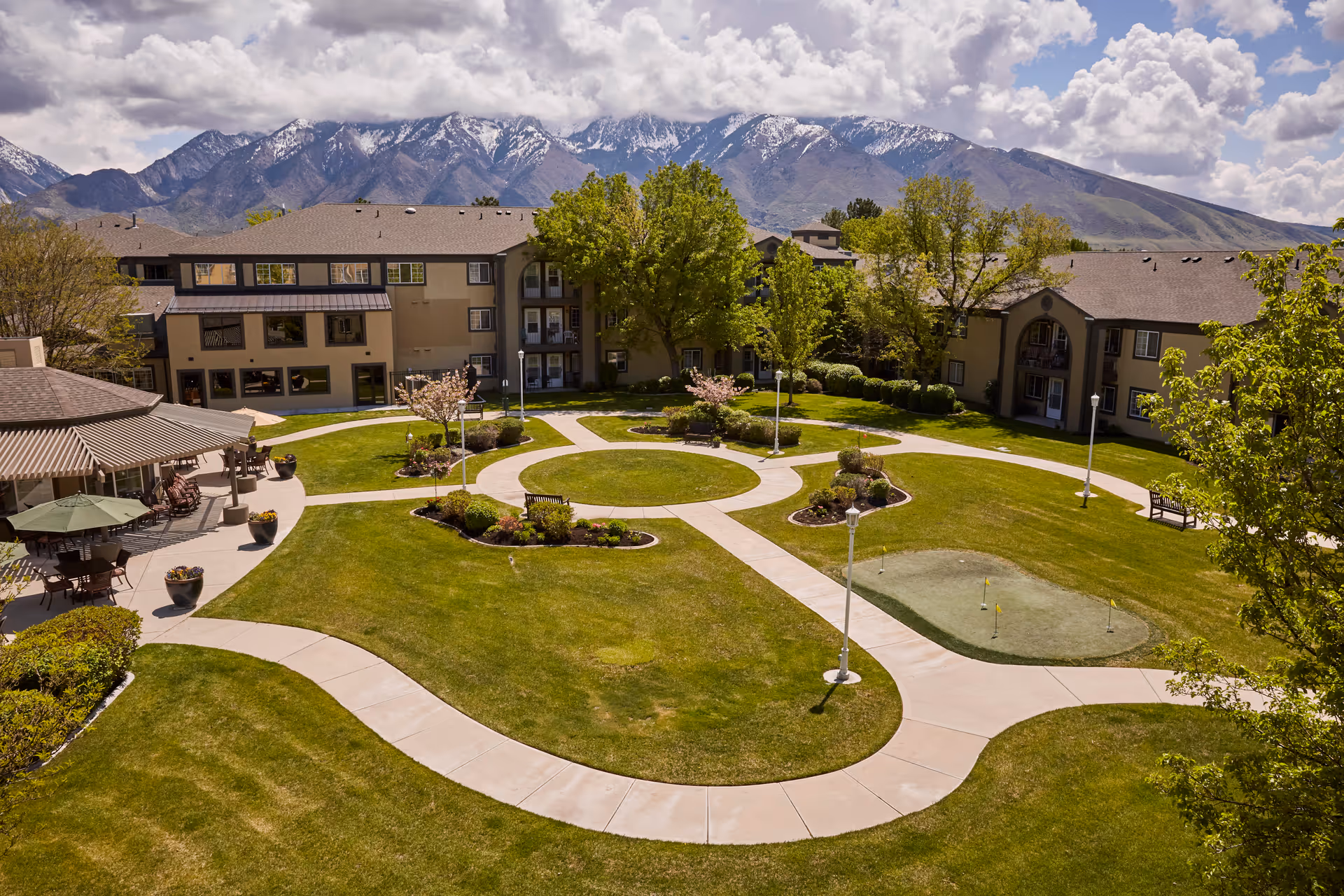 A landscaped outdoor courtyard area of a senior living facility with green lawns, walking paths, benches, a small putting green, and a covered seating area with tables and chairs. The facility buildings surround the courtyard, and snow-capped mountains are visible in the background under a partly cloudy sky.
