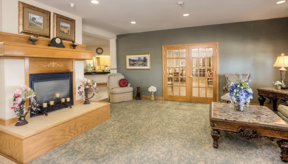 A cozy living room area with a wooden fireplace decorated with candles and floral arrangements. There are two armchairs, a wooden coffee table with a floral centerpiece, and a side table with a lamp. The room has a green accent wall with a framed picture and wooden-framed glass double doors leading to another room.
