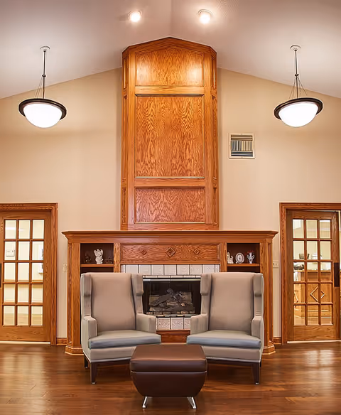 A cozy interior room featuring a wooden fireplace with built-in shelves on either side, two gray upholstered armchairs positioned in front of the fireplace, and a small brown ottoman between them. The room has wooden floors, beige walls, two pendant lights hanging from the ceiling, and two wooden-framed glass doors on either side of the fireplace.