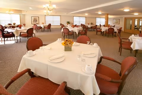 A spacious dining room with multiple tables covered in white tablecloths, each set with cups, plates, napkins, and glasses. The chairs have red upholstery and wooden armrests. The room is well-lit with ceiling lights and chandeliers, and there are large windows with blinds along the walls.