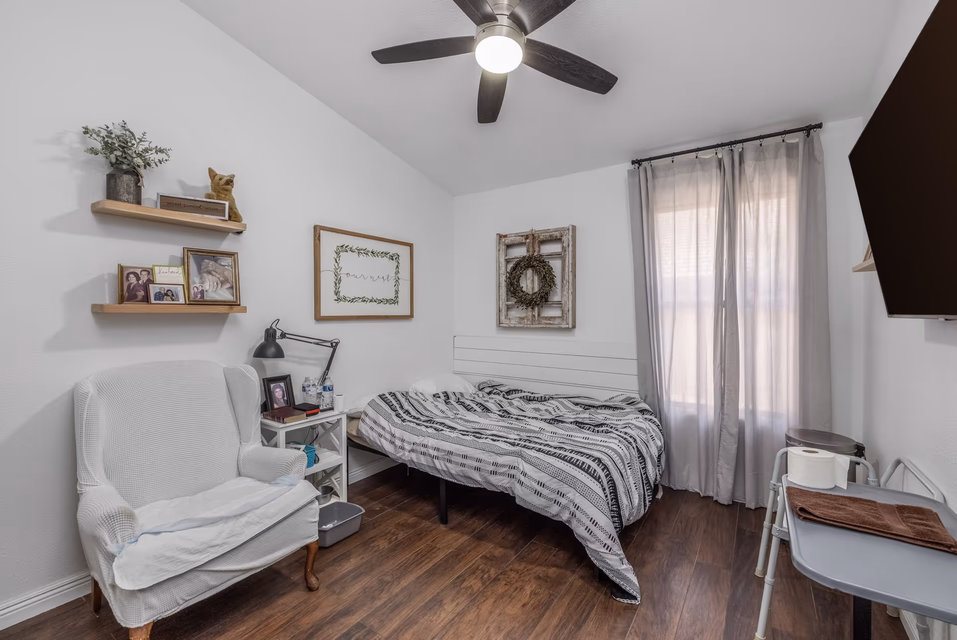 A cozy bedroom with a bed, armchair, wall shelves, framed decor, and a ceiling fan.