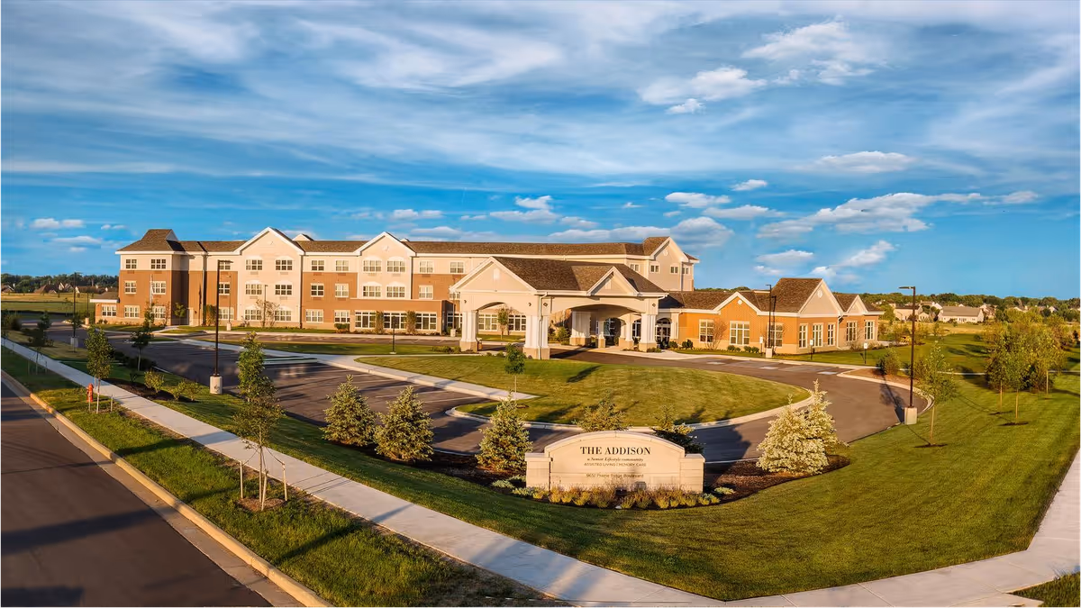 Front exterior of a large three-story retirement community building with a circular driveway, landscaped lawns, and a sign reading 'The Addison'.