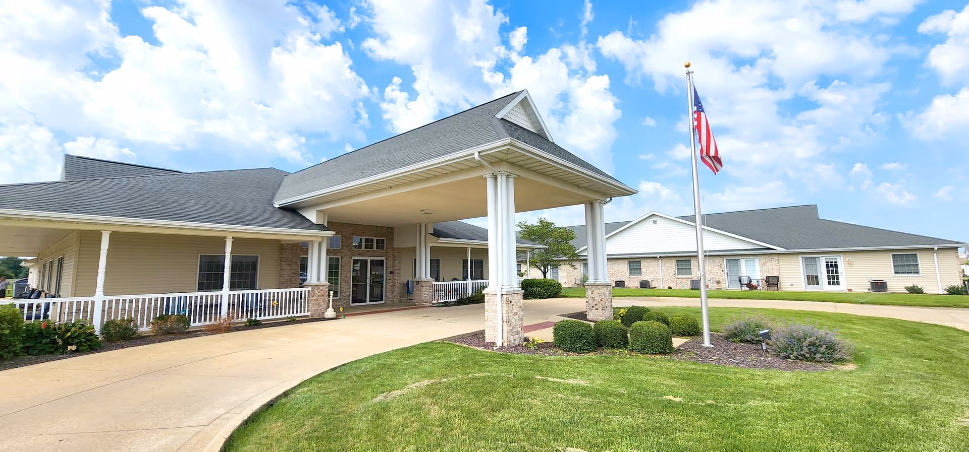 Exterior view of The Glenwood Supportive Living of Mt. Zion building with a covered entrance supported by white columns, a curved driveway, neatly trimmed bushes, a flagpole with an American flag, and a partly cloudy blue sky.