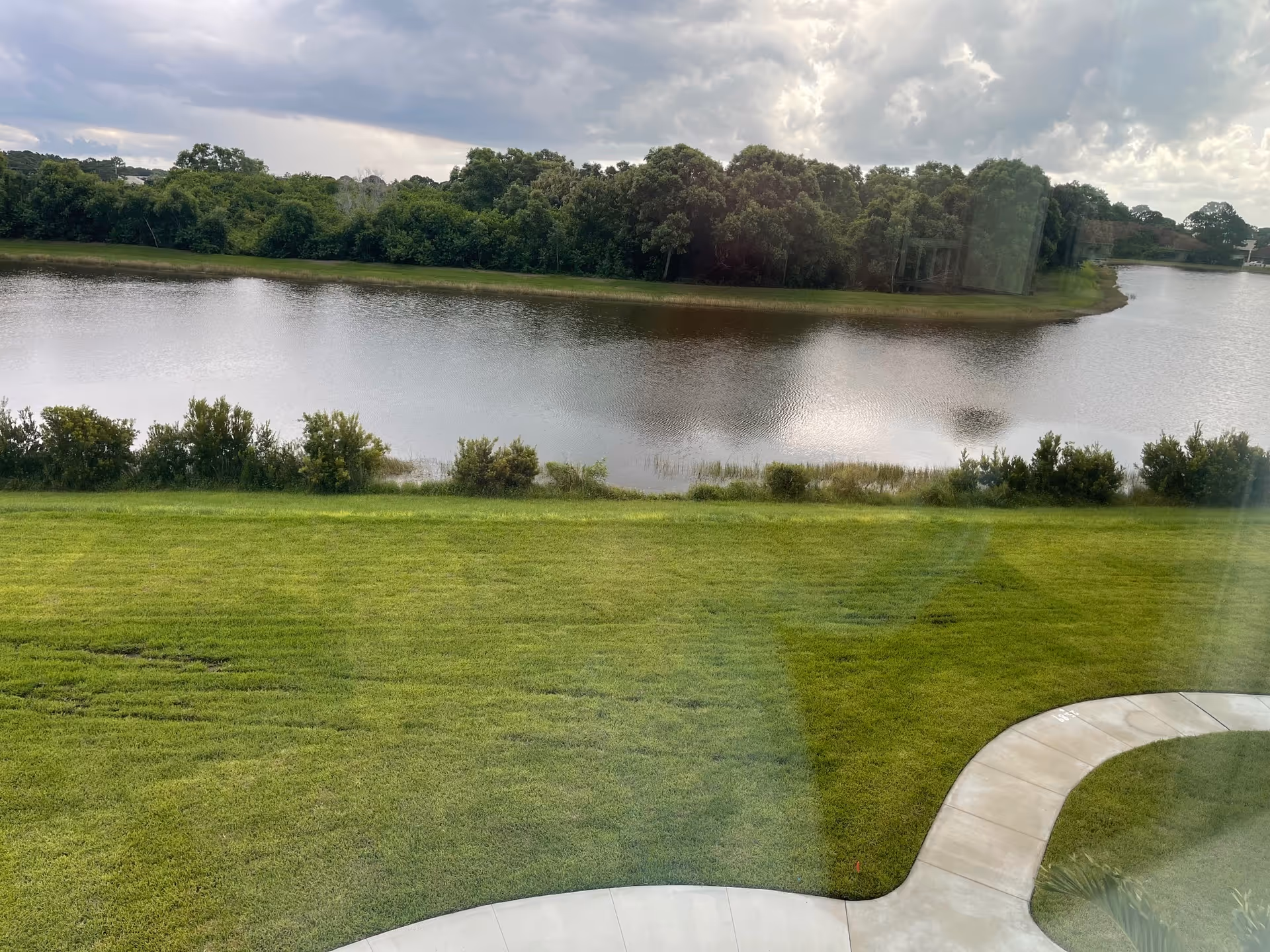 Grassy lawn with a curved sidewalk in the foreground overlooking a pond and tree-lined shore under a cloudy sky.