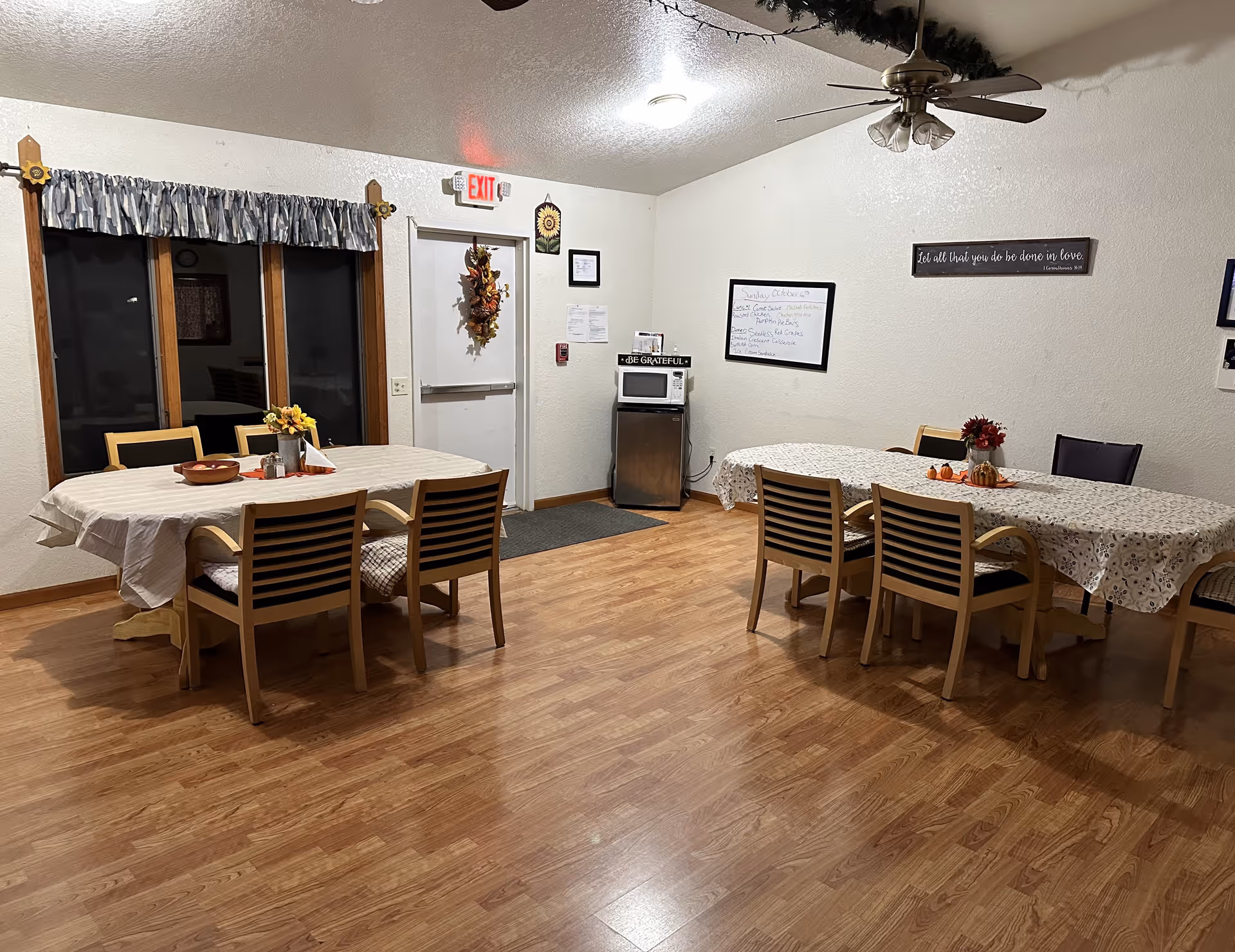 A dining room with two tables covered with tablecloths and surrounded by wooden chairs with black cushions. The room has wood flooring, a ceiling fan with lights, a window with a valance, and a door decorated with a fall wreath. There is a small microwave on a stand against the wall, a whiteboard with notes, and a sign that reads 'Let all that you do be done in love'.