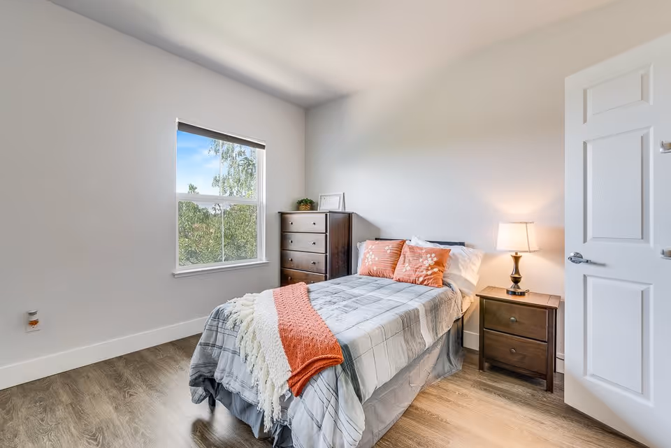 A bright and clean bedroom with a single bed covered in a gray and white plaid comforter, two orange decorative pillows, and a white and orange throw blanket. Next to the bed is a wooden nightstand with a lamp, and against the wall is a tall wooden dresser. A window shows greenery outside, and the room has light-colored walls and wood flooring.