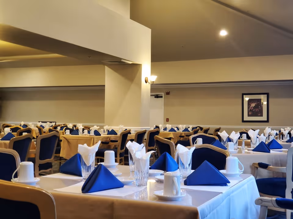 A dining room set up with multiple tables covered in white tablecloths and beige overlays, each table neatly arranged with blue and white folded napkins, clear drinking glasses, white coffee cups on saucers, and silverware. The room has beige walls, a framed picture on the far wall, and soft lighting from wall sconces and ceiling lights.