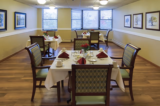 Dining room with several tables set for meals, upholstered chairs, red napkins, and a bay of windows.